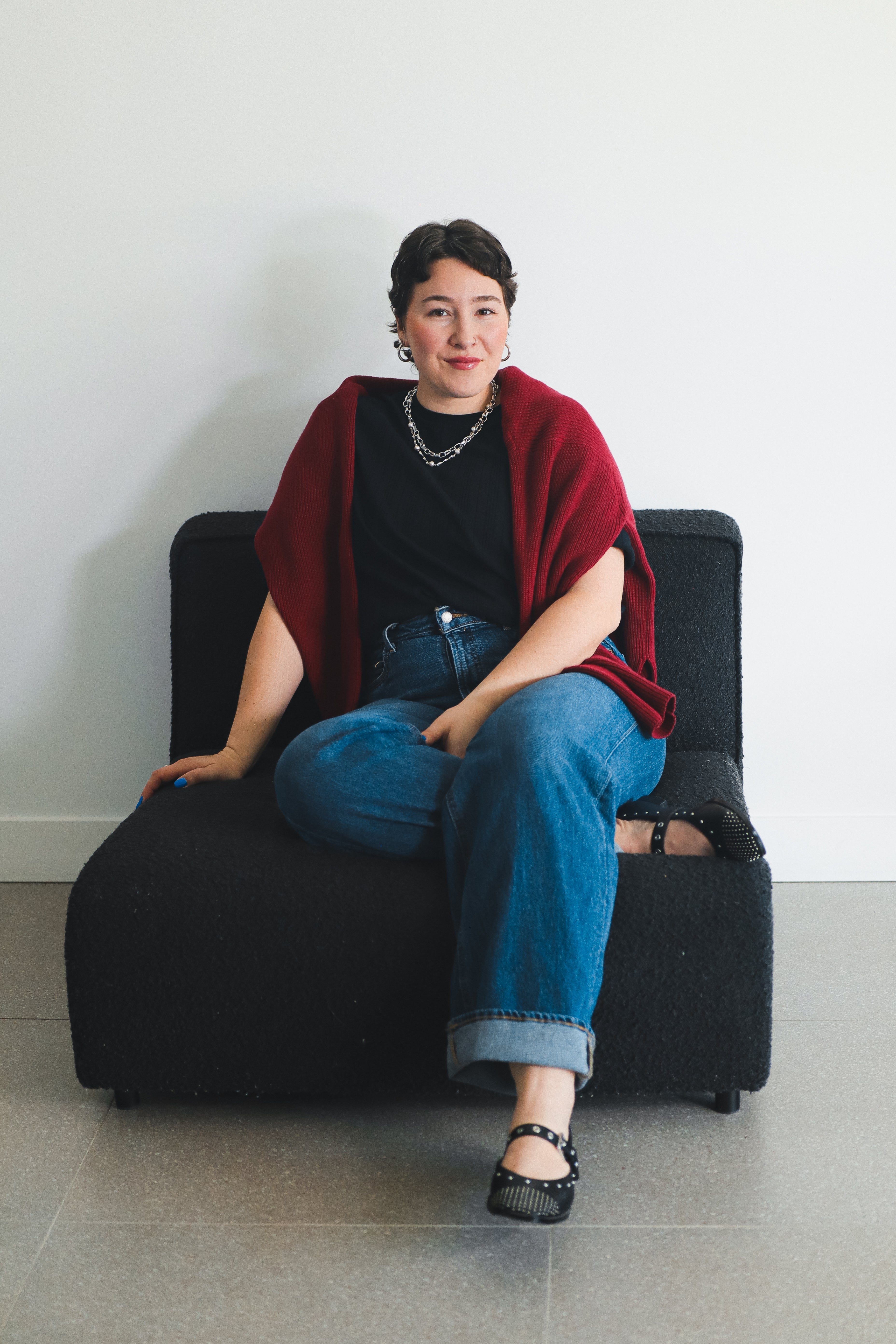 Person sitting on a black chair wearing a red sweater, black top, blue jeans, and black shoes.