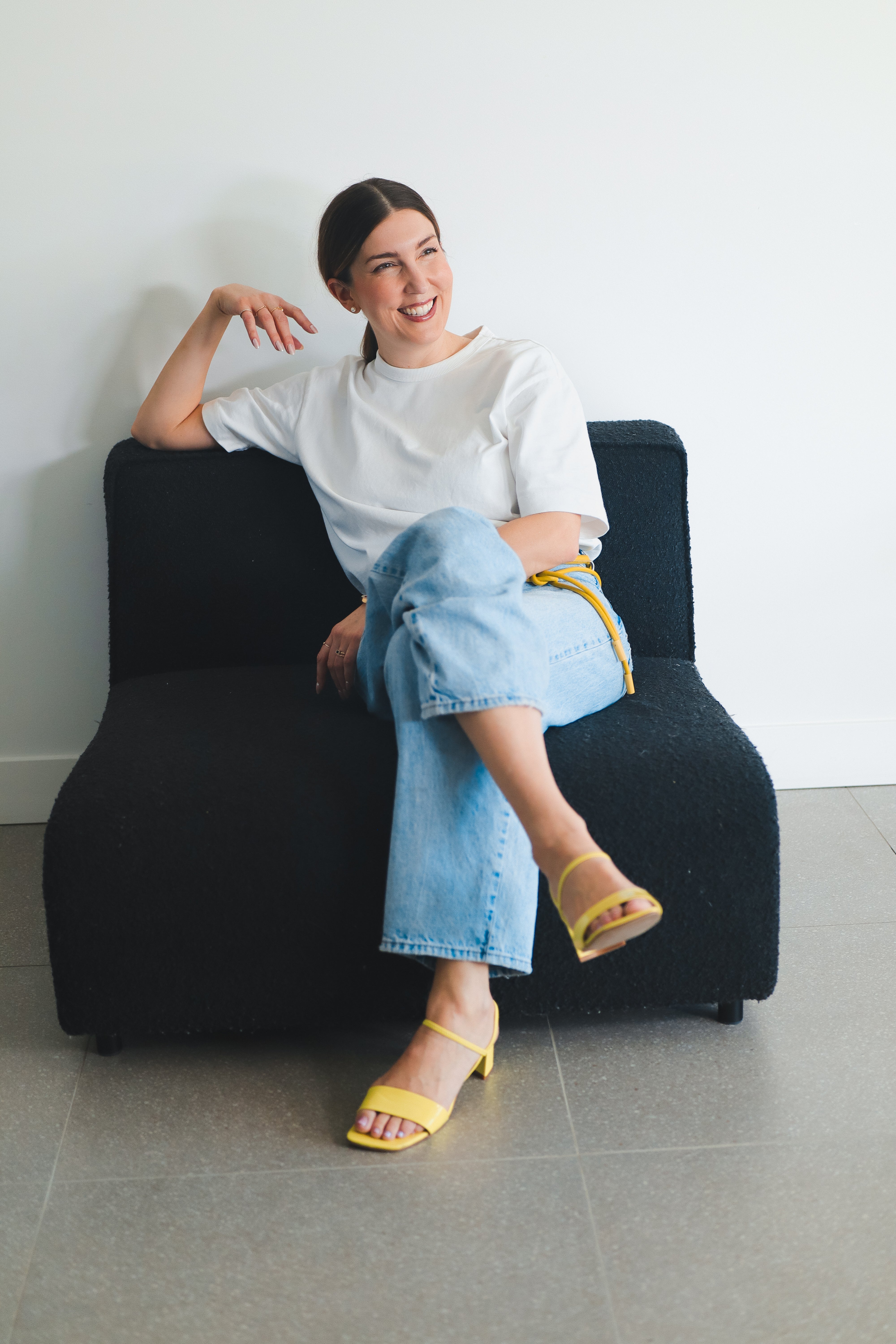 Woman sitting on a black chair wearing a white shirt, blue jeans, and yellow sandals.